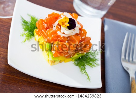 Fish tartare with raw salmon and diced avocado in shape of colorful cube decorated with pansy on white plate
