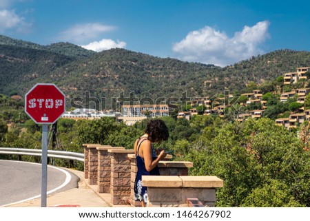 Long shot of a woman watching her mobile phone next to a particular stop traffic signal. In the background green mountains. Girona, Spain