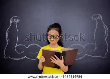 Picture of nerdy schoolgirl reading a book while showing her biceps on a blackboard in classroom 