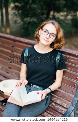 Young attractive woman with short haircut and glasses reads  book while sitting on bench outdoors