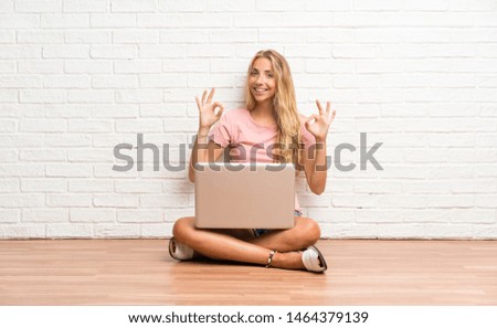 Young blonde student girl with a laptop on the floor showing an ok sign with fingers