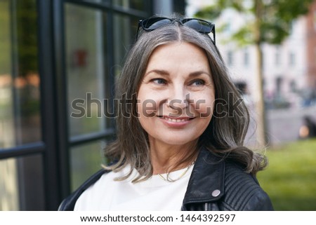 Modern urban lifestyle, city, people and mature age concept. Picture of happy joyful female pensioner wearing trendy shades and leather jacket posing outdoors, enjoying beautiful summer day