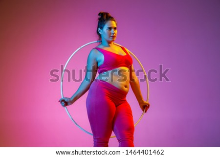 Young caucasian plus size female model's doing exercises on gradient purple background in neon light. Training with the hoop. Concept of sport, healthy lifestyle, body positive, equality.