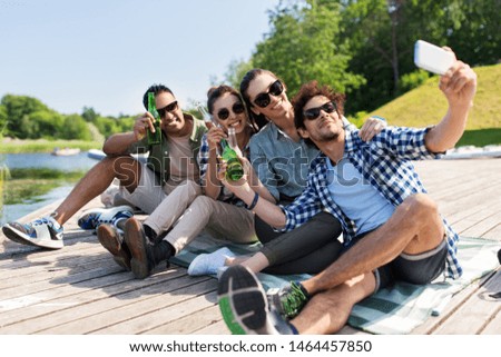 leisure, picnic and technology concept - friends with drinks taking selfie by smartphone on lake pier in summer park