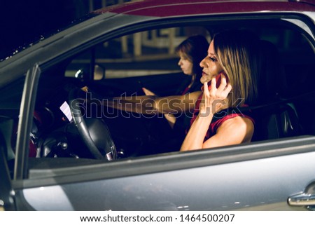 Two women female driving the car while holding mobile phone making call talking to the friends woman in the night