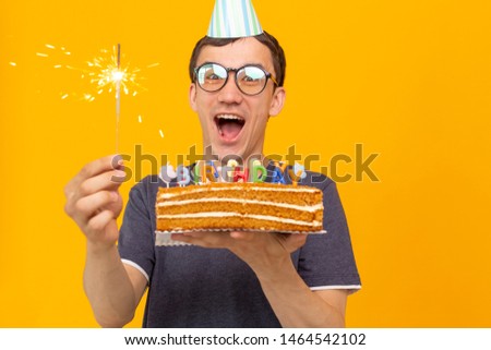 Funny positive guy in glasses holds in his hands a homemade cake with the inscription happy birthday posing on a yellow background. Concept of holidays and anniversaries.