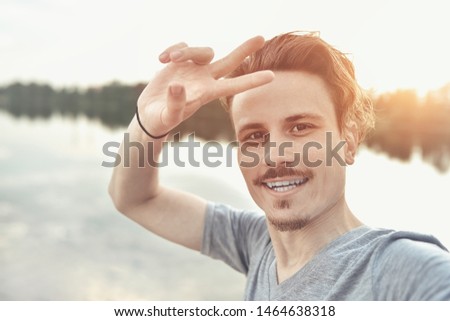 portrait of Young handsome stylish smiling guy makes selfie against the lake. peace sign