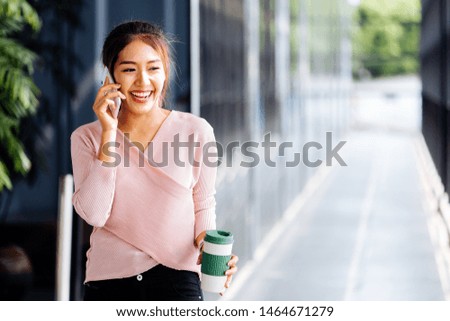 Young Asian woman talking on the mobile phone holding a coffee cup at office exterior background. Smart businesswoman discussing business talk outdoors