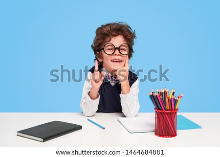 Cheerful little boy in nerdy glasses smiling and pointing up while sitting at desk against blue background during lesson at school