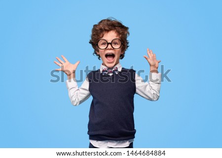 Little genius in nerdy glasses and school uniform yelling in excitement and holding hands up against blue background