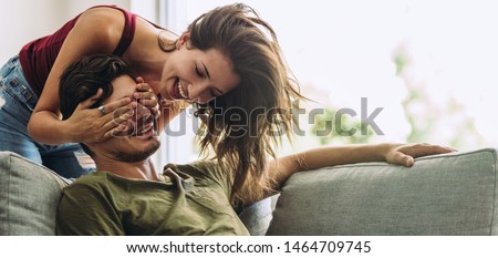 Smiling young woman covering her partner's eyes sitting on sofa. Young woman surprising man in the living room at home. Royalty-Free Stock Photo #1464709745
