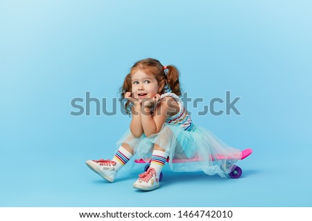 Cool little toddler girl in bright clothes sits on a skateboard and looking at the camera isolated on blue background