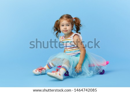 Cool little toddler girl in bright clothes sits on a skateboard and looking at the camera isolated on blue background