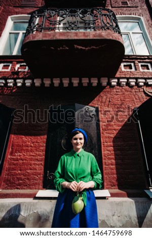 The concept of fashionable summer, blue and green colors. Stylish young woman in blue beret and skirt with a green shirt is standing against a red brick house close up