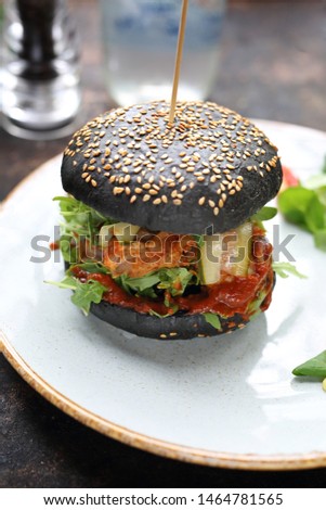   Burger in a black bun with chopped meat, pickled cucumber and ketchup served on a plate.