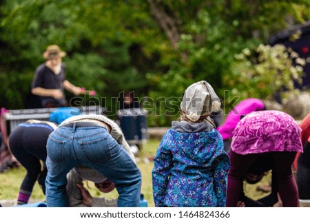 Fusion of cultural & modern music event. A young girl curiously watches adults doing yoga stretches, with heads bent to knees, a man playing glockenspiel is seen blurry in background, copy space above