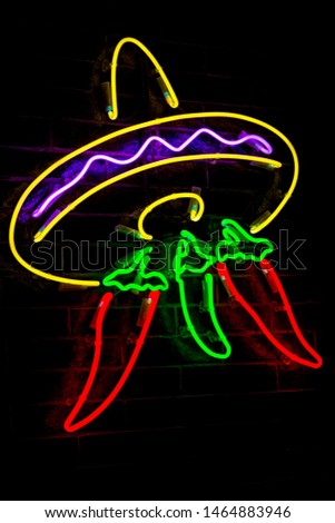 Glowing Neon stylized mexican sombrero and red peppers and blurred lights on black brick wall background. Dark tones vintage image.