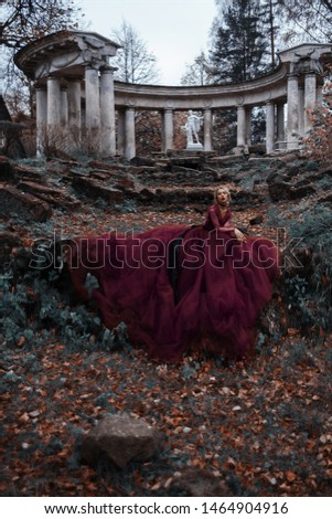 Young fragile girl in a red long dress on the background of the forest and ancient buildings with columns like a fairytale
