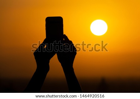 Silhouette of hands holding a mobile phone and making photography of the huge sun during sunset or sunrise. Male makes a photo on his phone