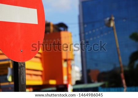 Traffic warning signs are erected at the entrance of the road, and the sun shines on the red one-way sign that is forbidden to enter, with a blurred yellow and blue building in the background.