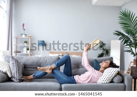 Beautiful smiling woman reading a book and lying on the sofa in the living room. Have a good time at home. Royalty-Free Stock Photo #1465041545