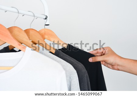 man's hand choosing black color t-shirt from collection of black, gray and white color t-shirt hanging on wooden clothes hanger in closet or clothing rack over white background