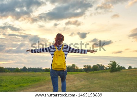 Young girl with yellow backpack, her back with open hands, nature background meadow dramatic sky, evening sunset sky