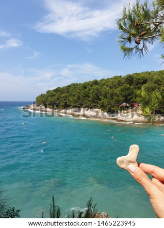 Woman fingers holding white stone heart with adriatic turquoise sea, green pine forest and blue sky on background. Vertical shot, copy space