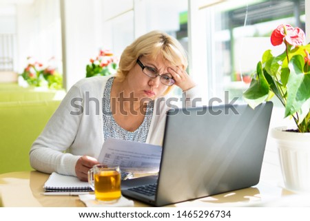 A middle-aged woman with a laptop works in a cafe in the office, she is a freelancer. A woman with glasses sits at a table with a Cup of herbal tea. She is puzzled.