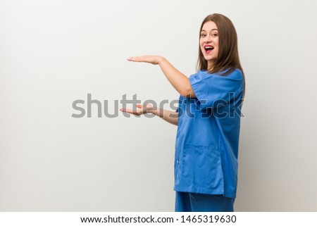 Young nurse woman against a white wall shocked and amazed holding a copy space between hands.