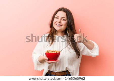 Young plus size curvy woman holding a cereals bowl smiling and raising thumb up