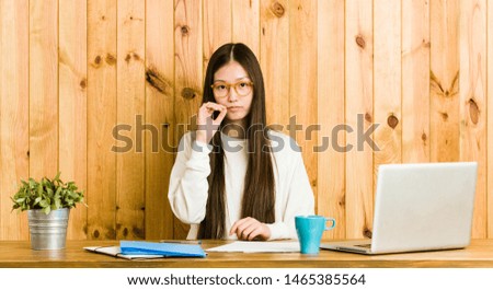 Young chinese woman studying on her desk with fingers on lips keeping a secret.
