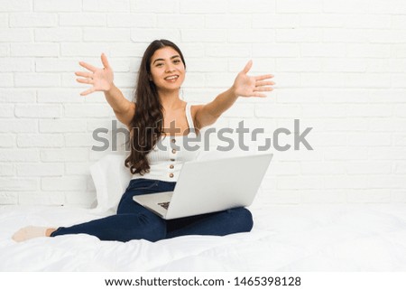 Young arab woman working with her laptop on the bed feels confident giving a hug to the camera.
