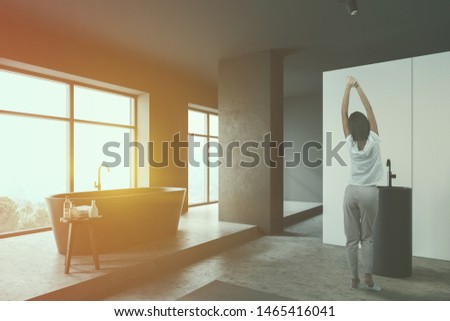 Young woman standing in stylish bathroom corner with white walls, concrete floor, comfortable gray bathtub, round sink and chair with towels and bottles. Toned image