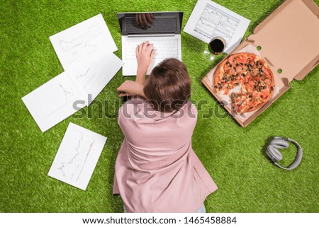 Woman having lunch with pizza and glass of wine working  on the grass using laptop with graphics and charts printed on the paper.