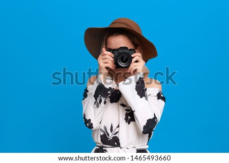 Portrait of a girl with curly blond hair in a sundown hat and white dress stands on a blue background. Model takes photos with retro camera.