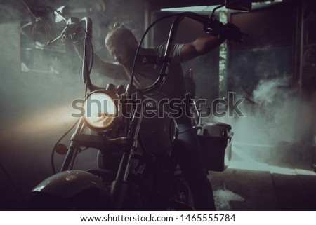 Handsome brutal man with a beard sitting on a motorcycle in his garage.