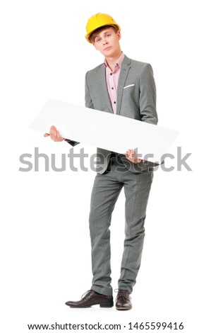 Full length portrait of young man in yellow safety helmet, holding empty board. Happy male with hard hat peeking from behind blank panel, isolated on white background. Guy showing blank placard board