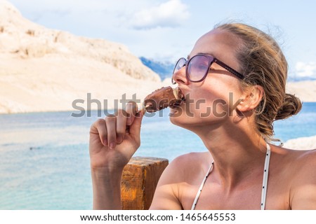 Beautiful caucasian girl eating ice cream on a stick in shade on picture perfect beach on Croatian Pag island in summer. Caucasian young woman model.