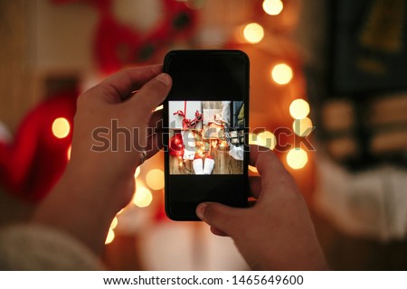 Hands holding phone and taking photo of christmas gift boxes, santa hat, lights on wooden background in dark room. Stylish hipster girl in sweater making christmas instagram flat lay photo