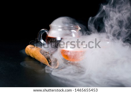 Glass tea cup, glass jug of tea, black tea in a scoop with vapour on a black background.