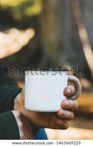 Dirty man's hand holding white enamel cup of hot beverage by an outdoor campfire with a vintage folk edit. Selective focus on mug with blurred background.