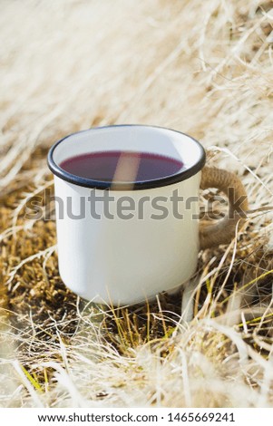 White enamel cup of hot beverage sitting on an dry grass with a vintage folk edit. Selective focus on mug with blurred background.