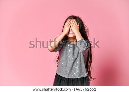 Happy brunette little girl in a dark dress covering her face with both hands on a pink background Royalty-Free Stock Photo #1465762652