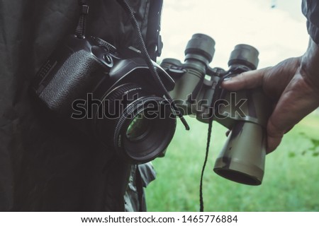 Male hand is holding binoculars on the background of nature; the camera is hanging near