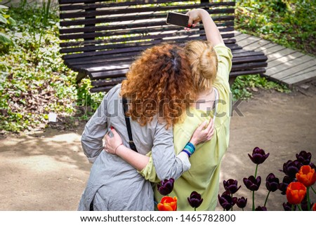 Two girlfriends take pictures of themselves with a smartphone outdoors on a Sunny summer day against the background of tulips. Rear view