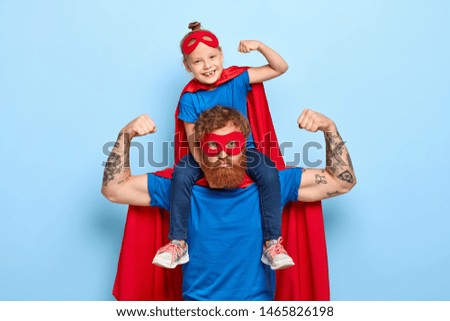 Strong powerful dad and little female child on his shoulders show muscles, ready to defend you and struggle, dressed in superhero clothes, isolated on blue background, prentend having superhuman power Royalty-Free Stock Photo #1465826198