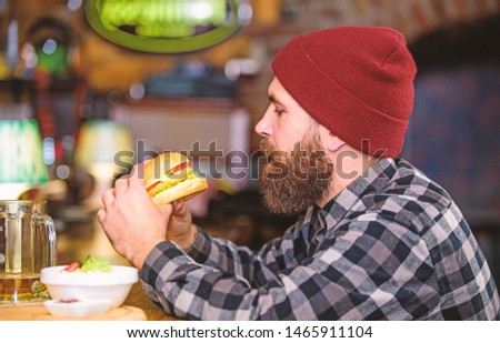 Pub is relaxing place to have drink and relax. Man with beard drink beer eat burger menu. Enjoy meal in pub. Brutal hipster bearded man sit at bar counter. High calorie snack. Hipster relaxing at pub.