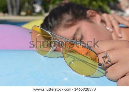 Young woman asleep,  sunbathing on a lilo in a swimming pool. Metal framed yellow sunglasses in foreground. Summer vibes, time off, fun and leisure