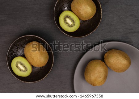 Group of four whole two halves of fresh green kiwifruit actinidia deliciosa in a dark ceramic bowl on a gray ceramic plate flatlay on grey stone